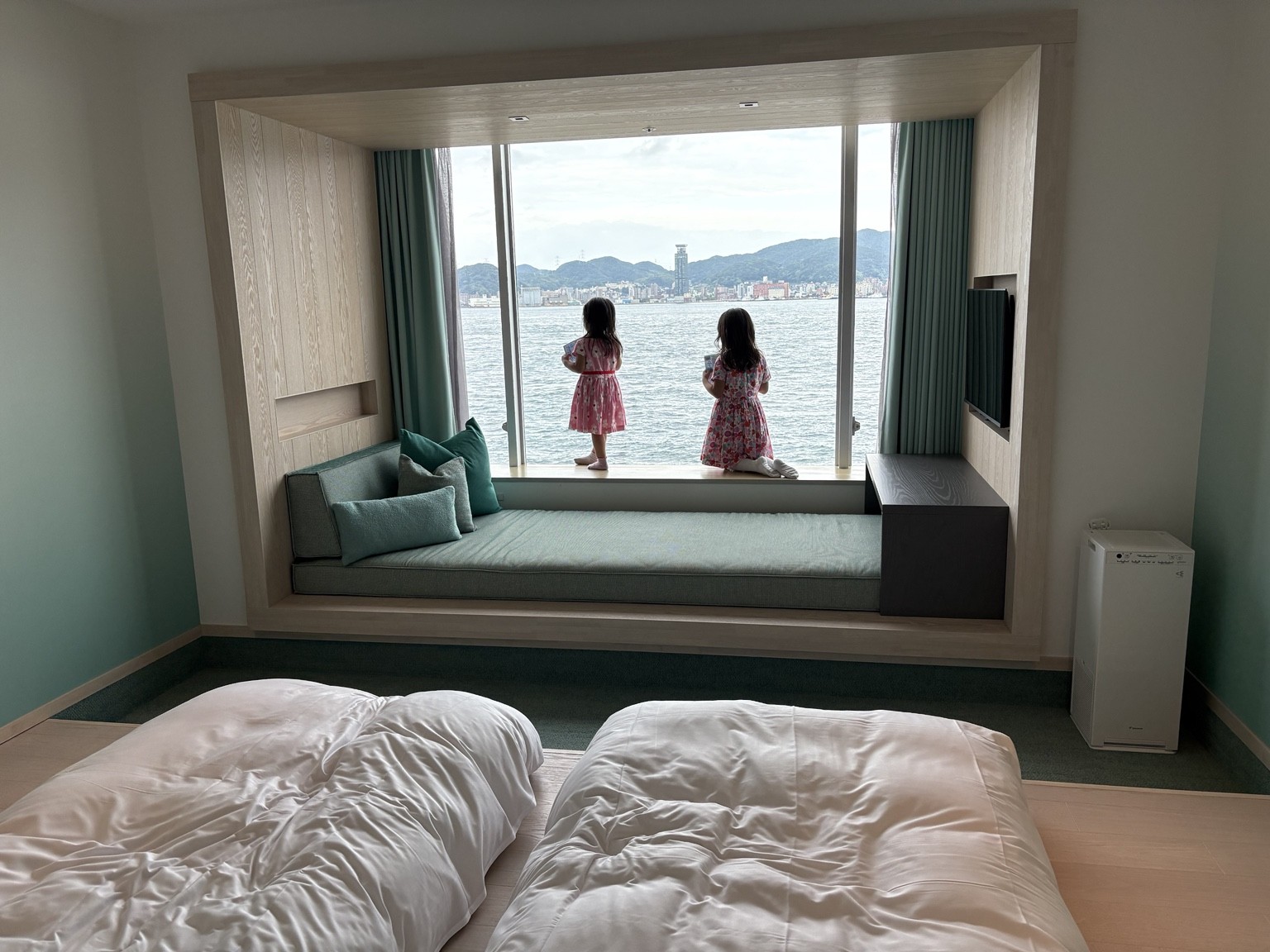 低いマットレスと窓辺のデイベッドがある客室で海を見る姉妹