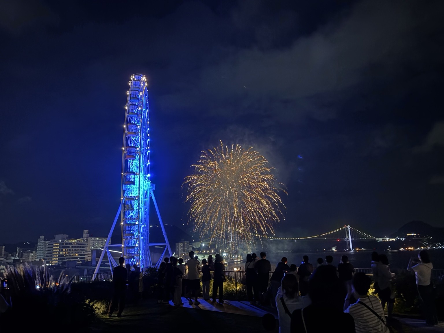 青く光る観覧車の奥に上がる花火と海峡の夜景
